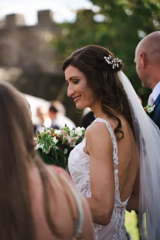 A smiling bride wearing an open-back lace dress and veil looks over her shoulder while holding a textured bouquet outdoors.
