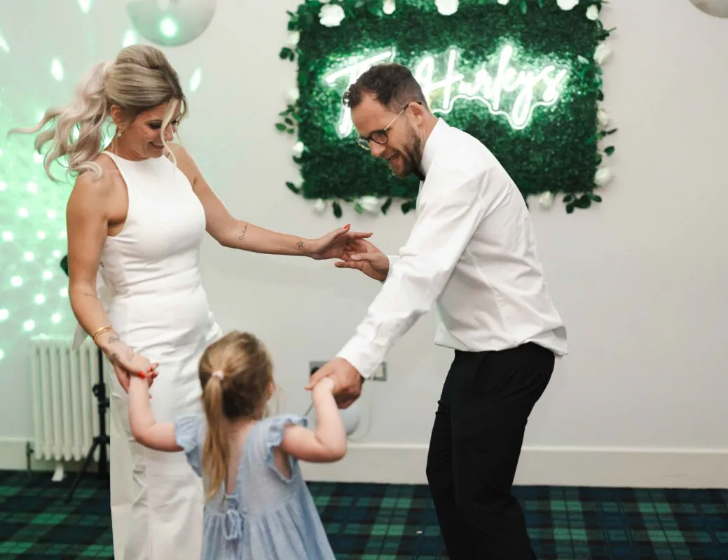 A bride in a white halterneck dress and a groom with rolled-up sleeves dance happily in a reception room while holding hands with a young child under a glowing neon sign.