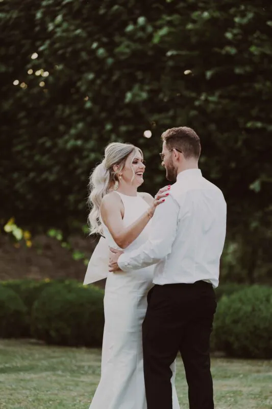 A newlywed couple shares a joyful laughing moment together outdoors against a lush green backdrop during their portrait session.