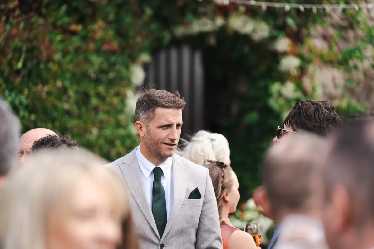 A groom in a light grey suit and dark green tie mingles with guests outdoors near an ivy-covered stone archway.