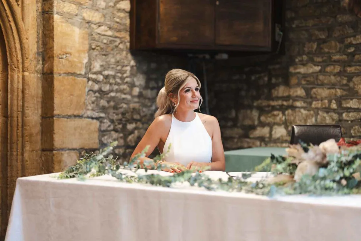 A smiling bride seated at a clothed table decorated with a greenery runner inside a historic stone venue during the wedding ceremony.
