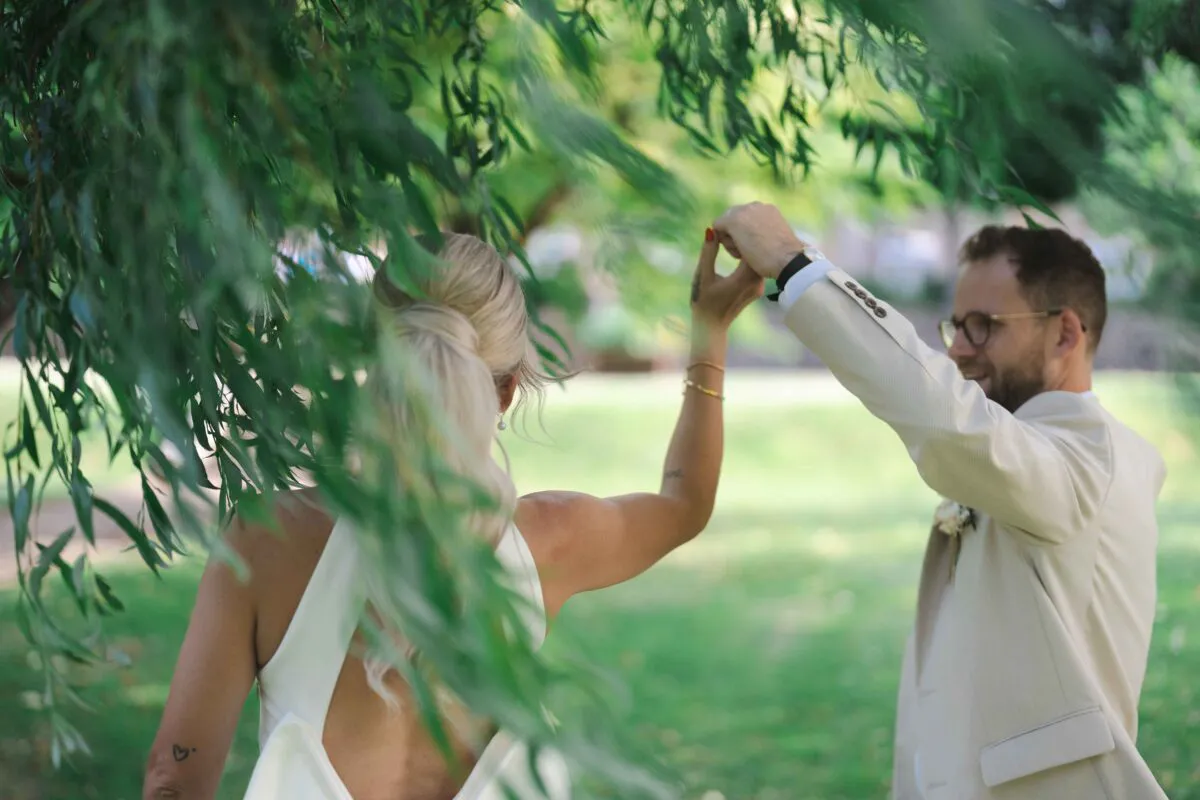 A newlywed couple holds hands outdoors while framed through the lush hanging branches of a weeping willow tree.