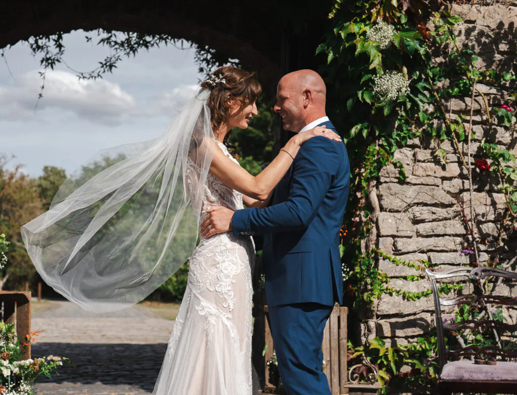 A newlywed couple embraces lovingly outdoors against an ivy-covered stone wall, with the bride's veil catching dramatically in the wind.