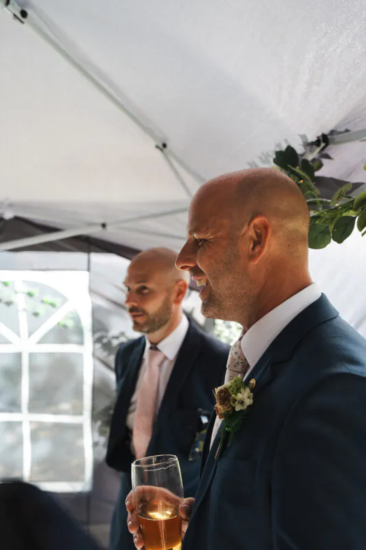Two smiling groomsmen in navy suits and pink ties holding drinks beneath a white marquee tent during a wedding reception.