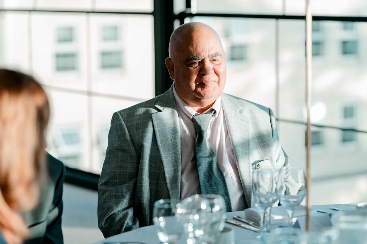 An older male wedding guest wearing a grey patterned suit and tie sits attentively at a reception table bathed in natural window light.