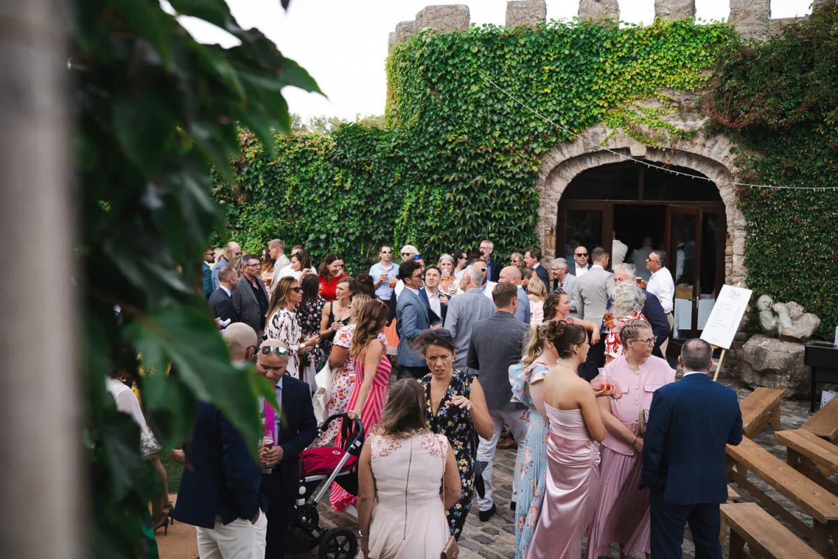 Wedding guests mingle during a lively drinks reception in a cobbled courtyard framed by an ivy-clad stone castle wall.