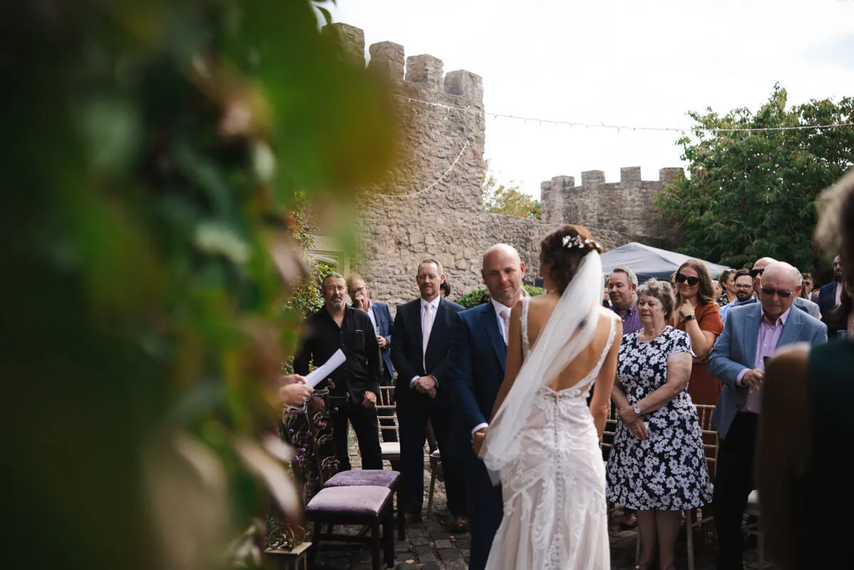 A bride and groom stand facing each other during an outdoor castle ceremony, framed by foliage in the foreground and watched by assembled guests.