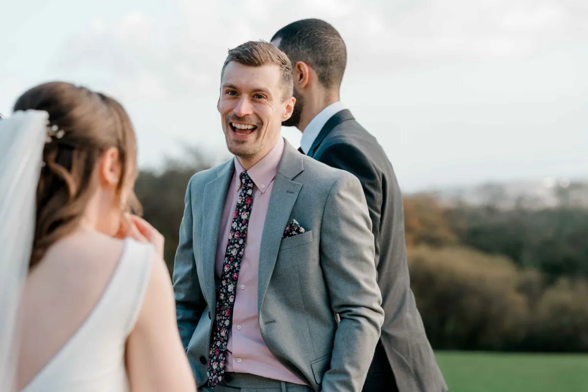 A groom in a grey suit and floral tie laughs happily while speaking with the bride outdoors during the wedding celebration.