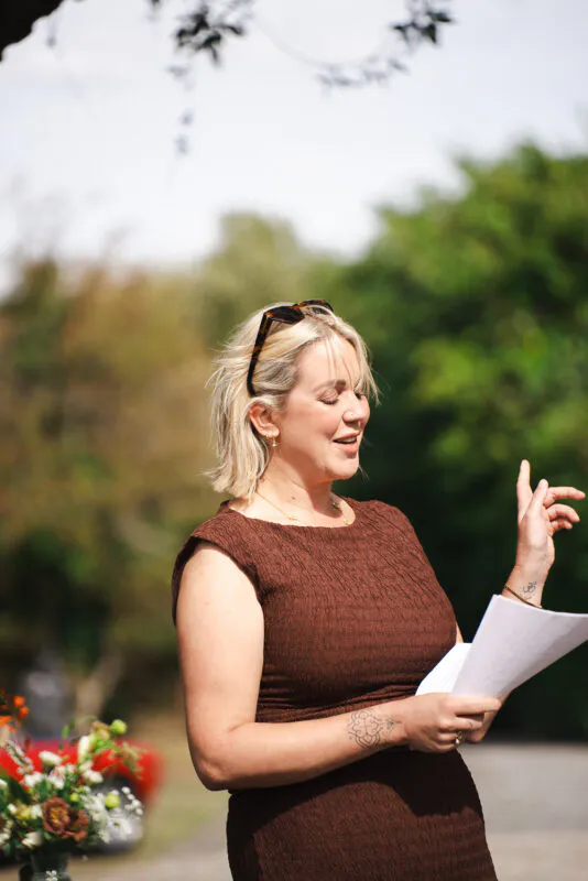 A blonde guest with tattoos and sunglasses on her head holds a speech outdoors in natural light.