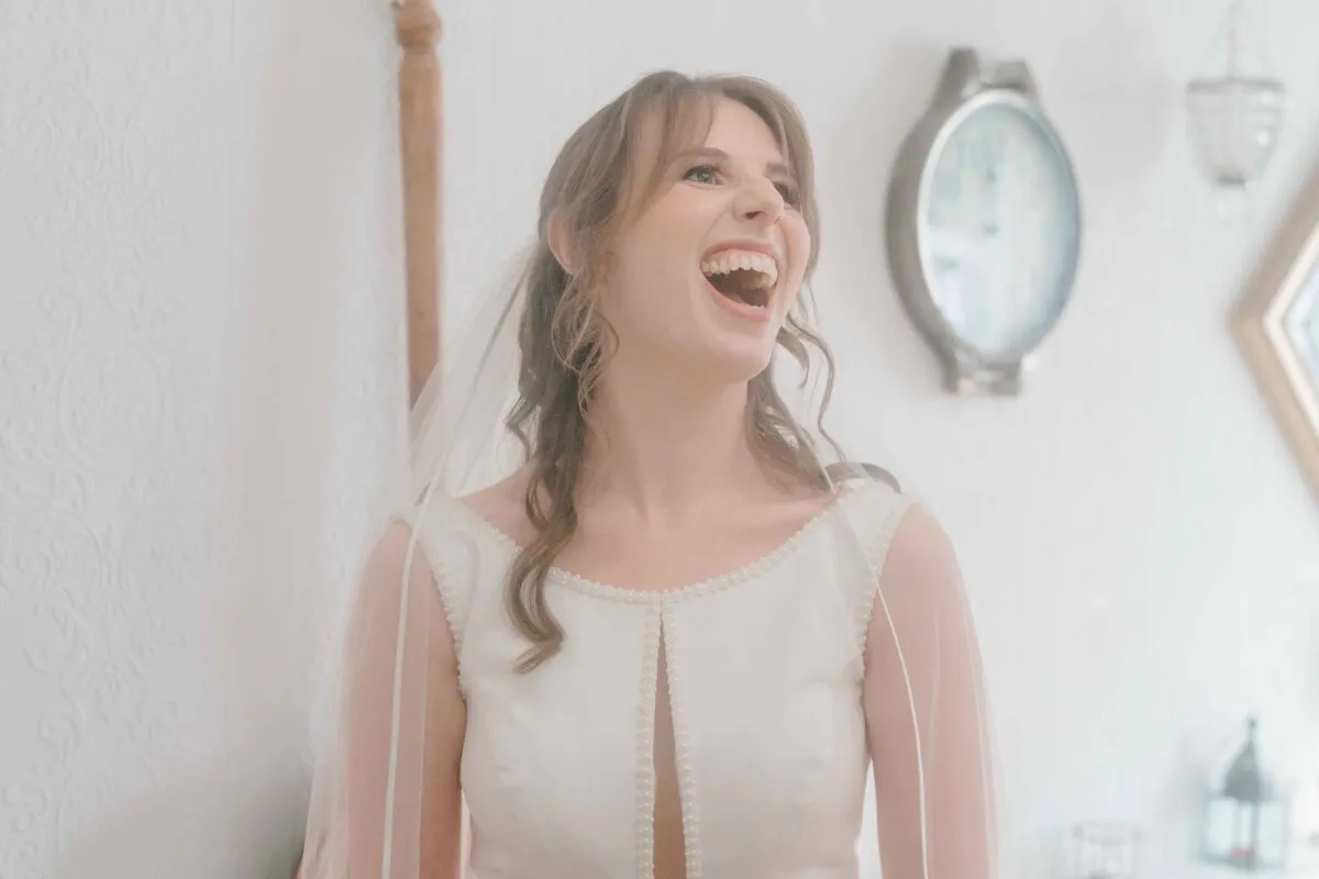 A joyful bride with soft wavy hair and a veil over the lens laughs heartily in a bright, light-filled room during wedding preparations.