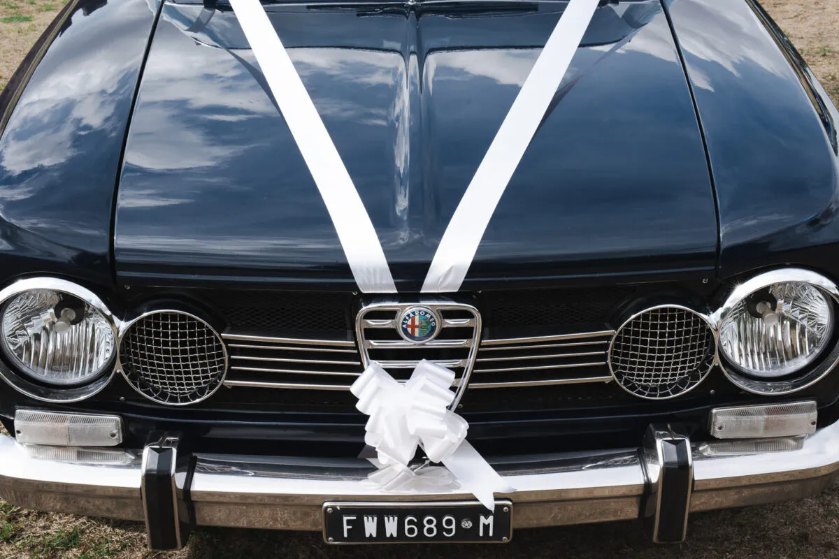 A close-up shot of the front of a classic dark wedding car adorned with elegant white ribbons and a bow across the bonnet.