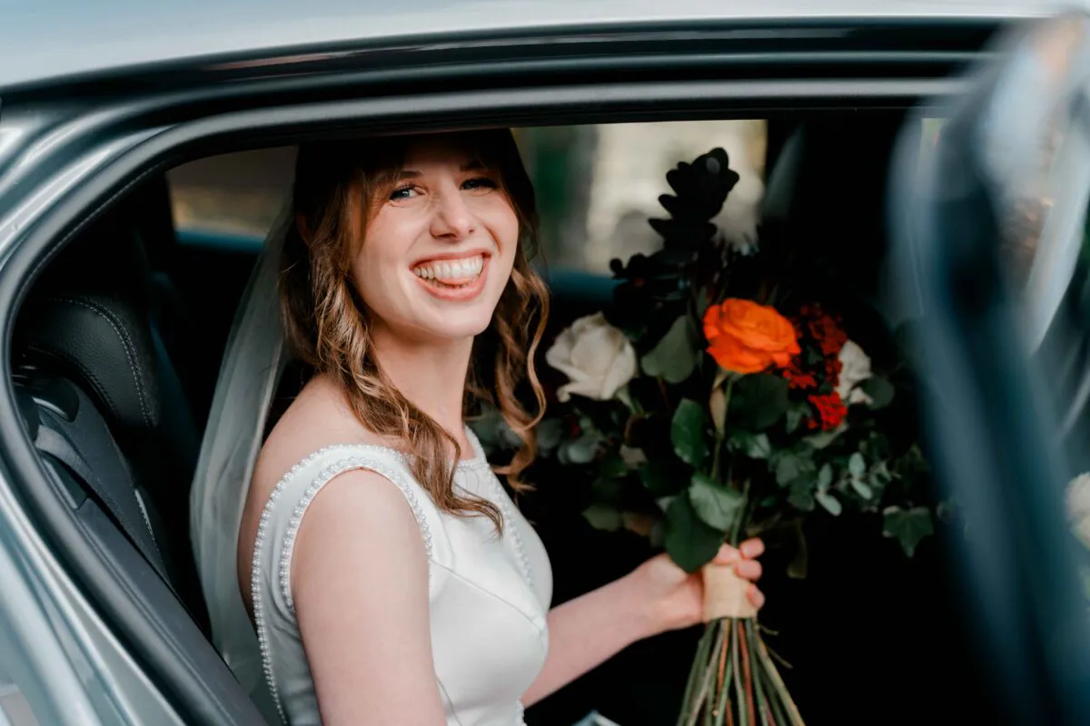 A smiling bride holding a vibrant rose and greenery bouquet looks out from the open door of a wedding car.