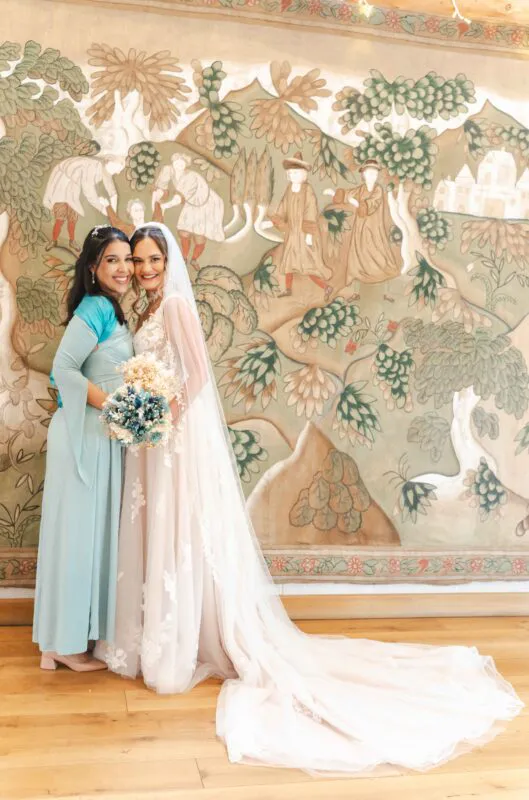 Two smiling brides stand side by side embracing in front of a large medieval-style tapestry, with one bride wearing a lace wedding dress with a long veil and holding a dried floral bouquet.