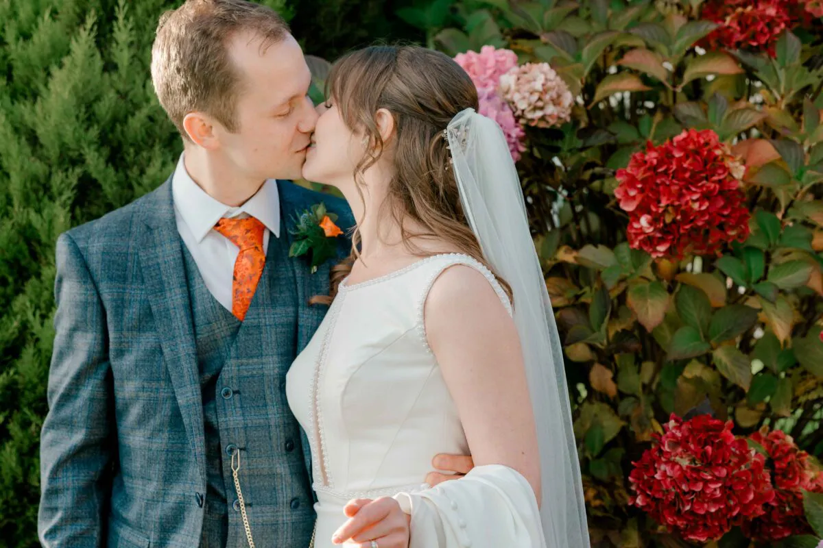 A newlywed couple shares a tender kiss outdoors against a vibrant backdrop of lush greenery and striking red floral blooms.