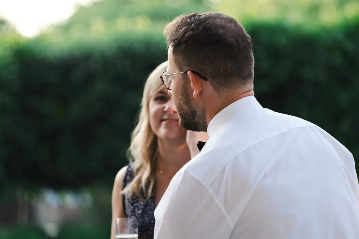 An over the shoulder shot of a groom in a white shirt looking towards a smiling wedding guest outdoors.