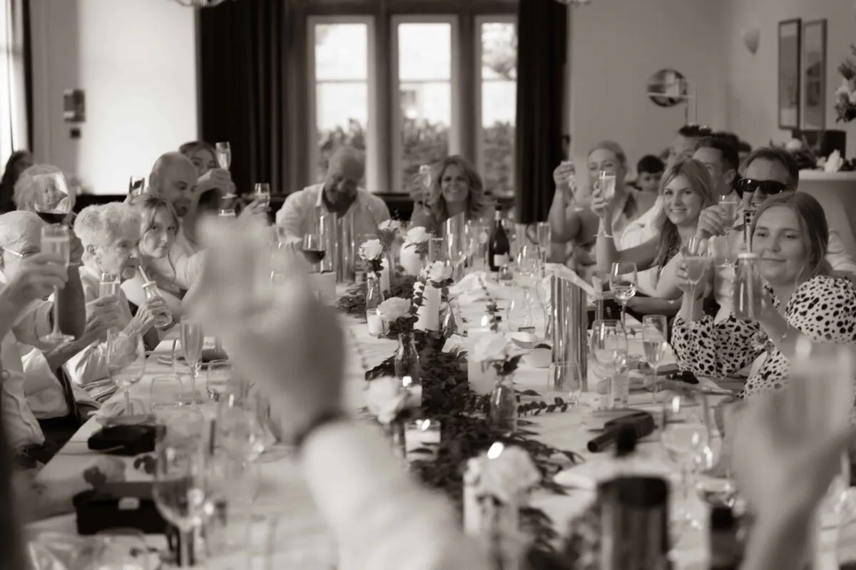 Wedding guests raise their glasses in a toast during the wedding breakfast, seated along a long banquet table decorated with candles and greenery.