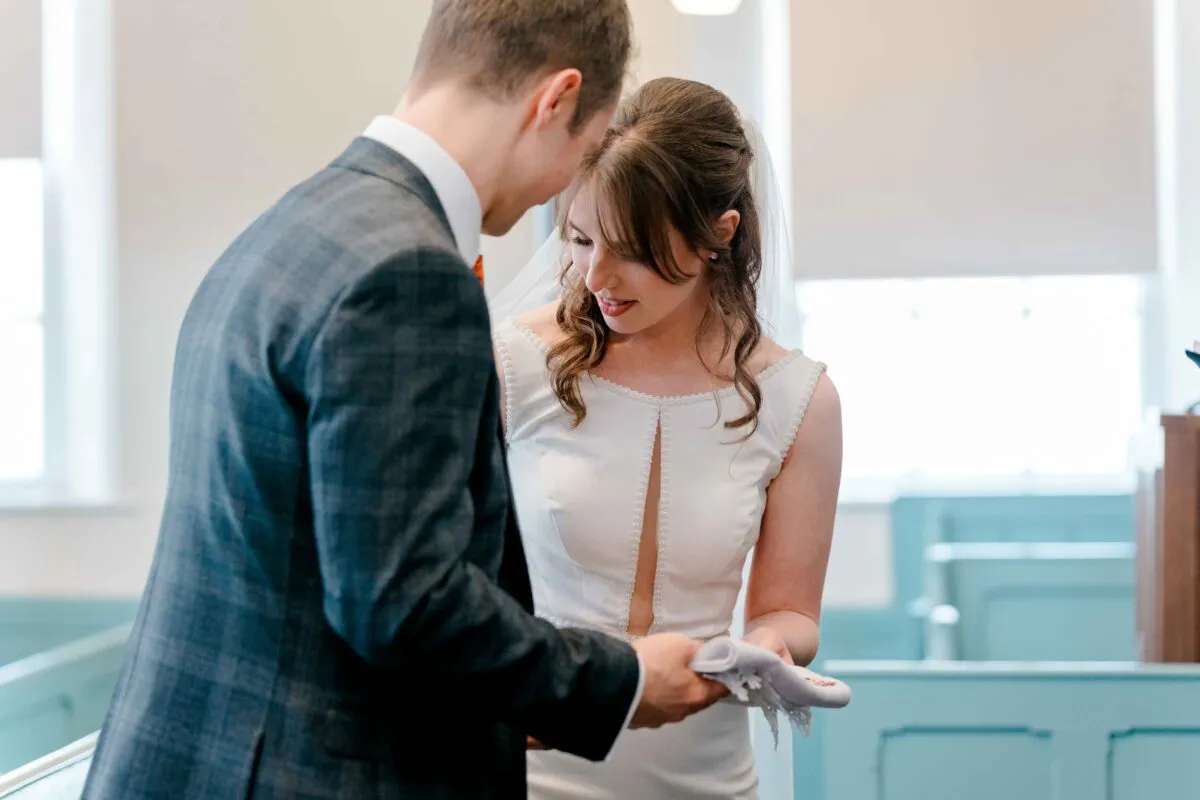 A groom in a tweed suit and a bride in a crepe gown with a square neckline look down affectionately during their church ceremony.