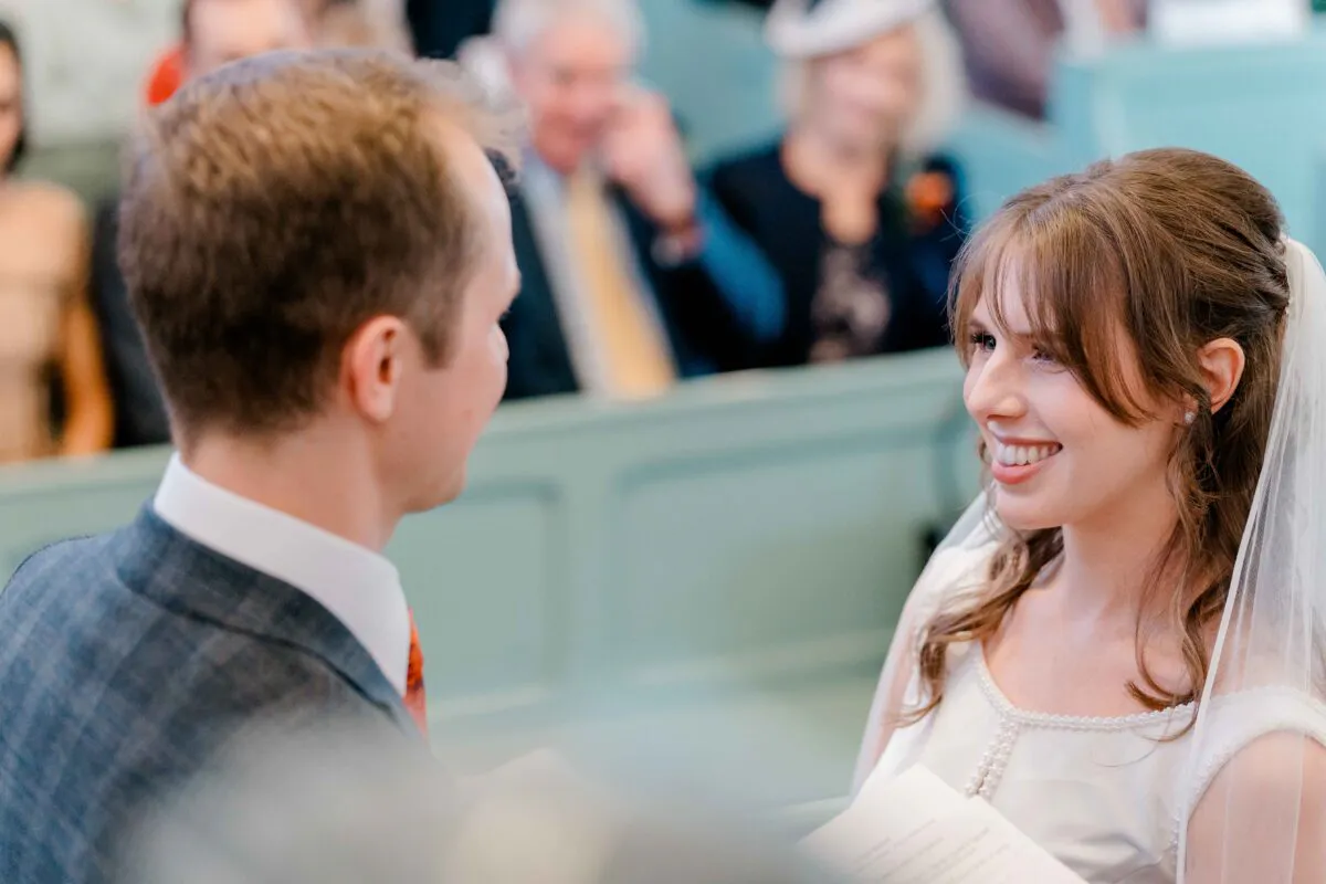 A smiling bride looks lovingly at her groom during the wedding ceremony inside a traditional church with seated guests watching on.