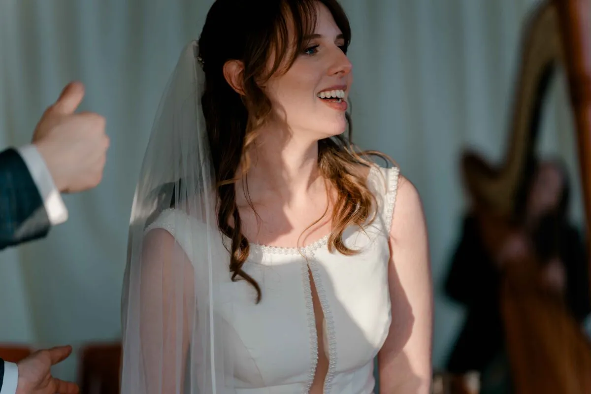 A smiling bride with half-up wavy hair and a pearl-trimmed gown laughs during a brightly lit wedding moment.