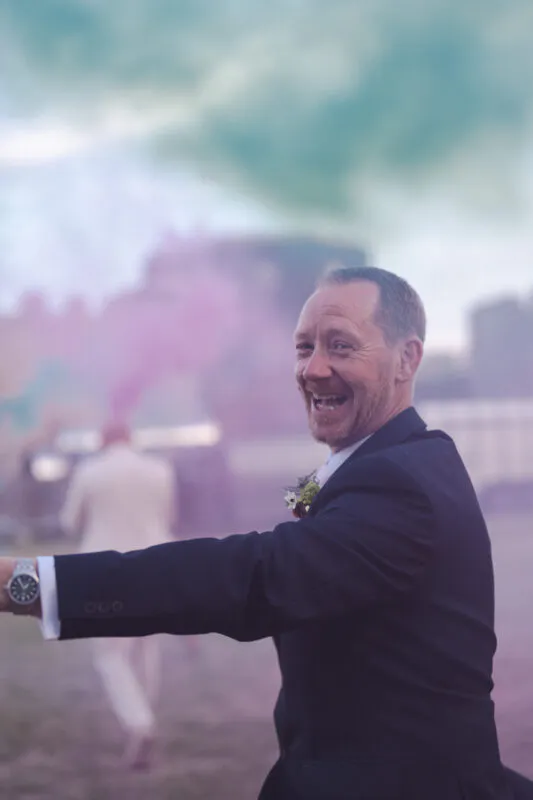 A groom in a black suit laughs joyfully outdoors, wearing a boutonniere and a watch, with a colourful hazy backdrop.