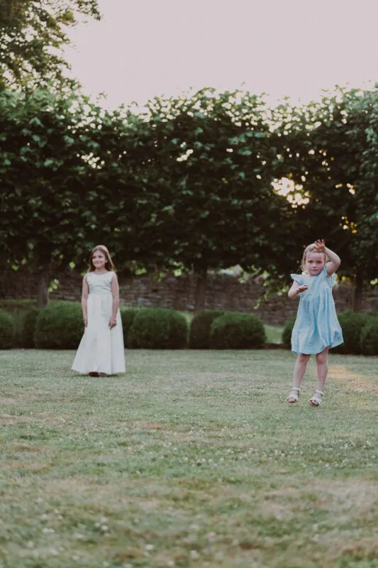 A young flower girl playfully jumps on a manicured lawn while a junior bridesmaid watches in the background against a backdrop of trees.