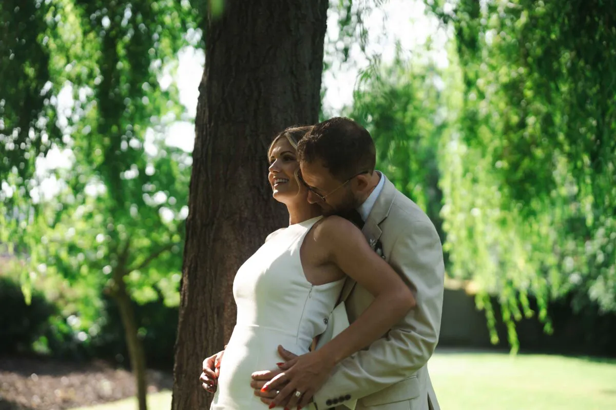 A groom gently embraces a smiling bride in a modern high neck gown against a lush green woodland backdrop.