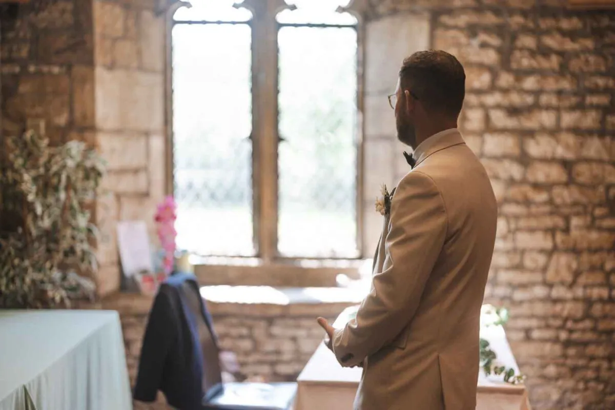 A groom wearing a beige suit and glasses stands inside a stone church, illuminated by soft window light as he awaits the ceremony.