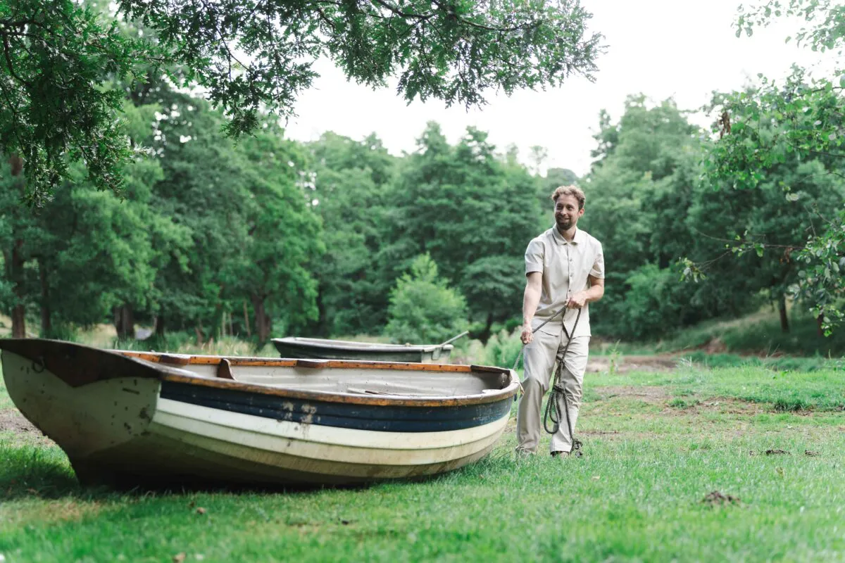 A groom in a casual beige suit pulls a traditional wooden rowing boat by a rope along a lush grassy lakeside surrounded by trees.