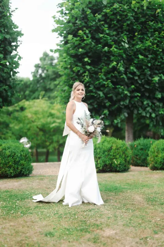A smiling bride holding a textured bouquet stands gracefully on a lush green lawn surrounded by manicured topiary and mature trees.