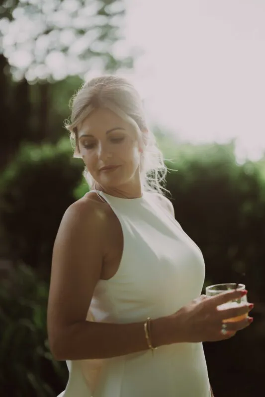 A graceful bride in a modern high-necked white gown holds a cocktail during an outdoor drinks reception, illuminated by warm backlight.