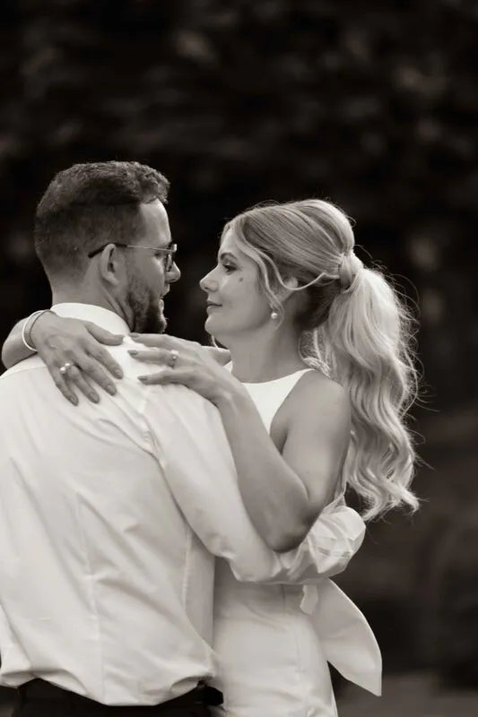 A newlywed couple shares an intimate first dance outdoors, captured in a warm sepia tone with the bride looking lovingly at the groom.