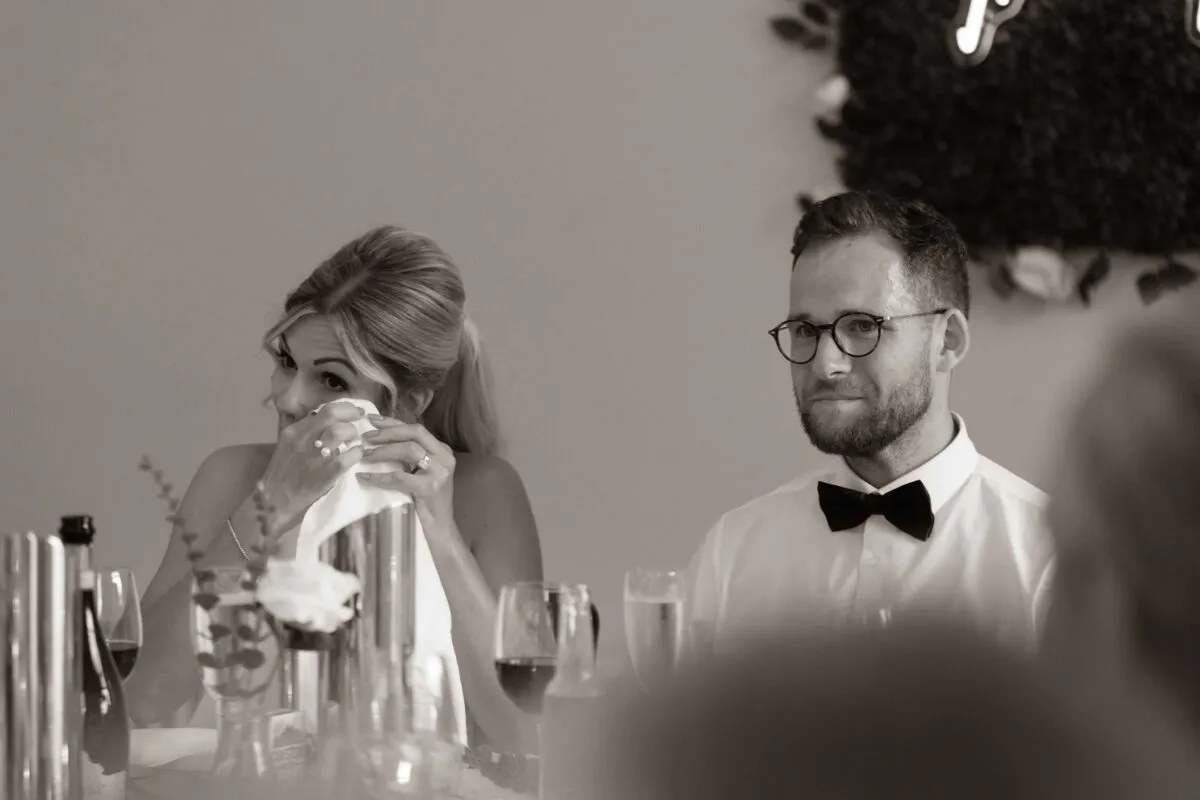 An emotional bride wipes a tear from her eye with a napkin during the wedding speeches while the groom looks on.