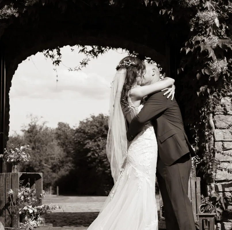 A newlywed couple shares a tender kiss framed within an ivy-covered stone archway in a classic black and white wedding portrait.
