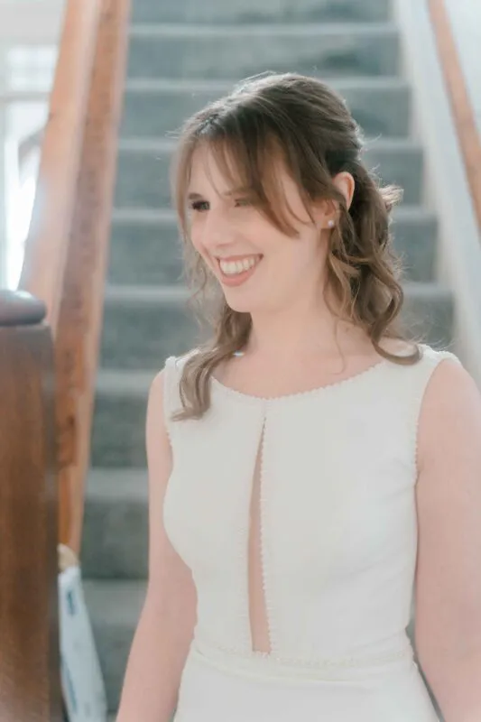 A smiling bride stands near a wooden staircase, wearing a white sleeveless gown with pearl embellishments and her hair styled half up.