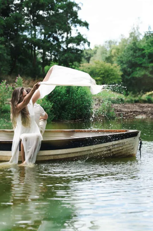A bride in a white slip dress playfully splashes water from a wooden boat on a serene lake while her partner sits beside her.