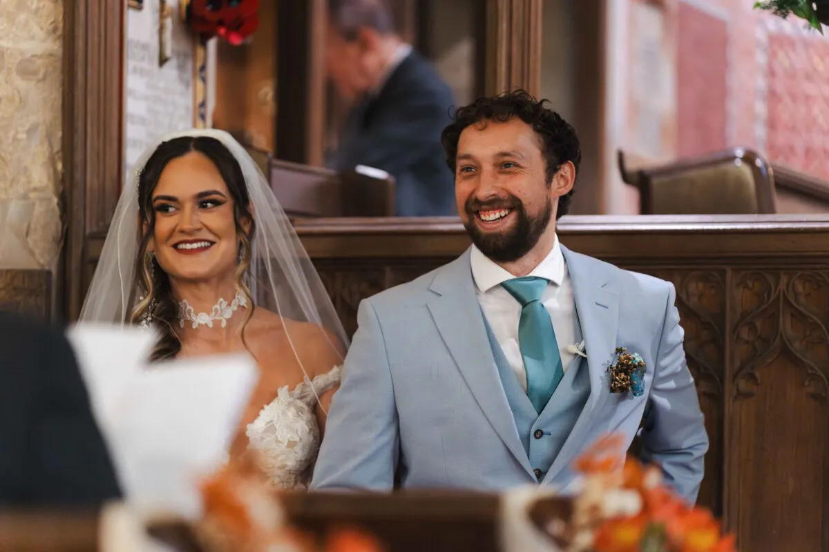 A smiling bride wearing a lace gown with a veil sits beside a groom in a light blue suit and teal tie during their wedding ceremony.