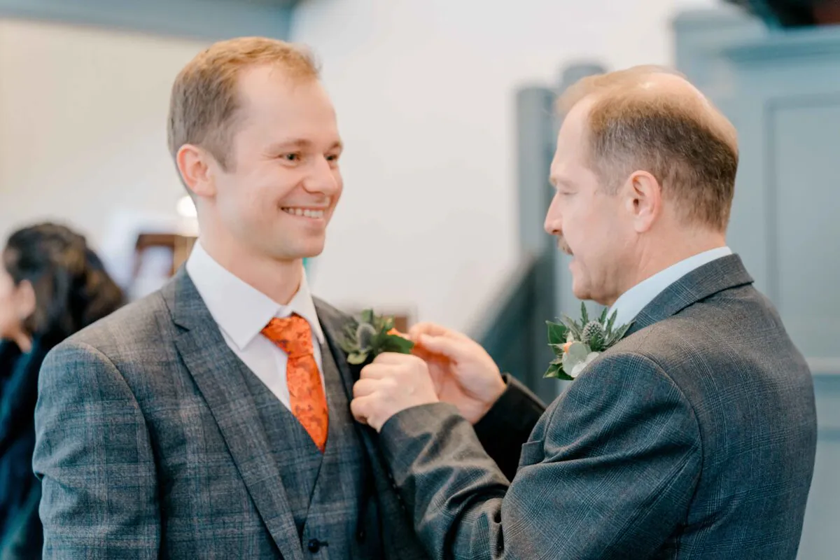 An older gentleman carefully pins a floral boutonniere onto the lapel of a smiling groom wearing a grey checked suit and orange tie.