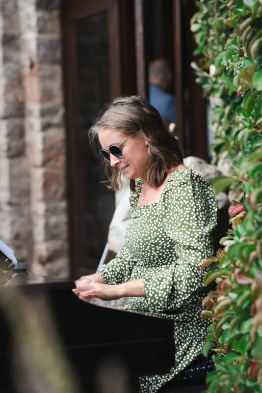 A guest wearing a green polka dot dress and sunglasses plays the piano outdoors near a stone wall and lush foliage.