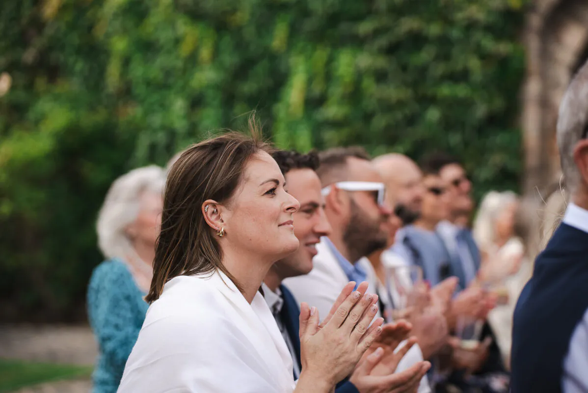 A smiling guest in a white top claps enthusiastically alongside other attendees seated outdoors in front of a lush ivy-covered wall.