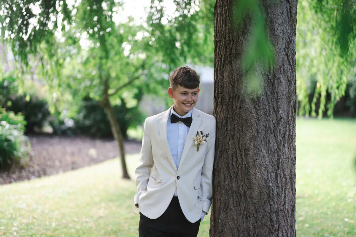 A young page boy wearing a cream suit jacket and a black bow tie smiles while leaning against a large tree in a lush garden setting.