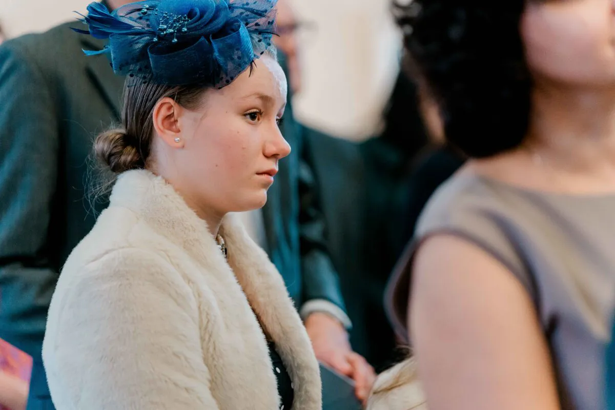 A young wedding guest wearing a blue fascinator and a cream faux fur stole looks attentively toward the ceremony.