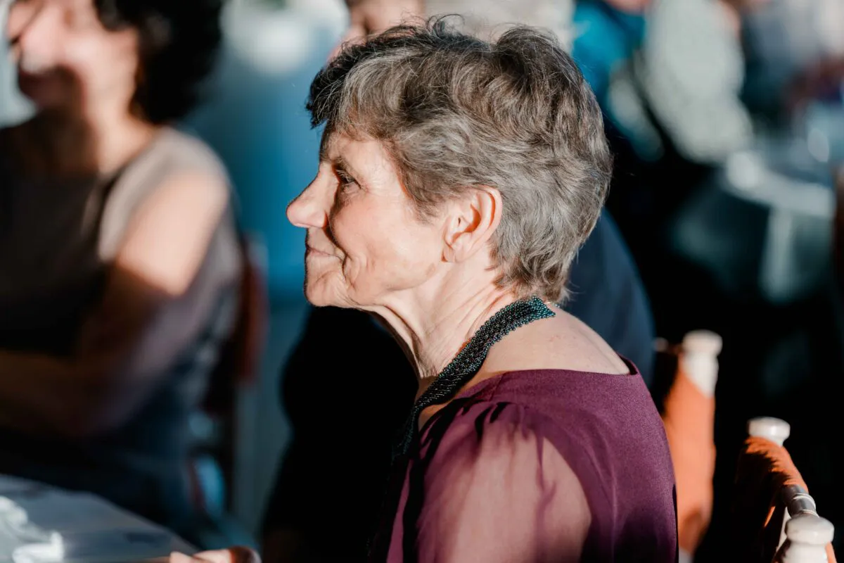 An elderly female wedding guest with short grey hair and a beaded necklace smiles warmly in profile under natural side lighting.