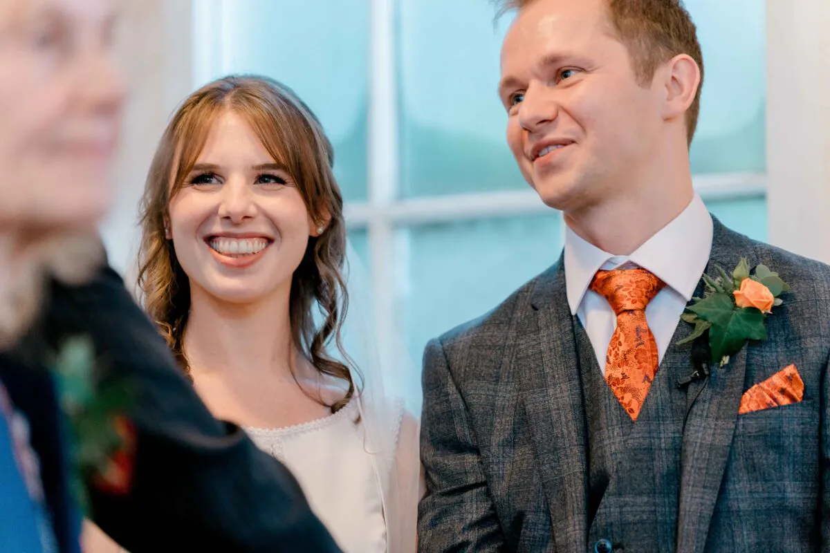 A joyful bride and groom smiling happily during their wedding ceremony, with the groom wearing a tweed suit and orange patterned tie.
