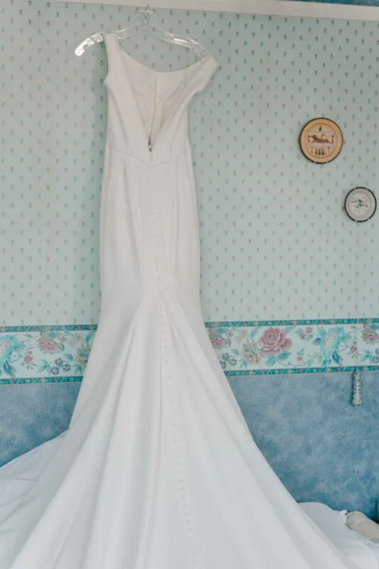 An elegant white crepe wedding dress hangs against patterned blue wallpaper, showcasing its fitted silhouette and long button-detailed back.