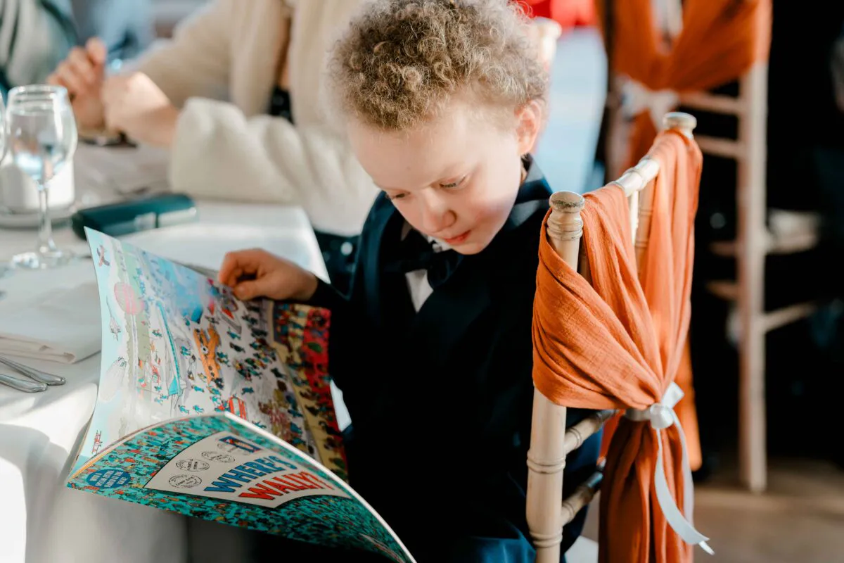 A young page boy sits quietly at a wedding reception table, engrossed in reading an illustrated activity book while wearing a smart suit.