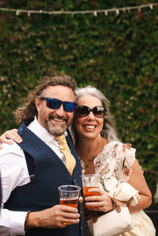 An older couple embracing and laughing together at an outdoor wedding reception, both wearing sunglasses and holding plastic cups of drinks.