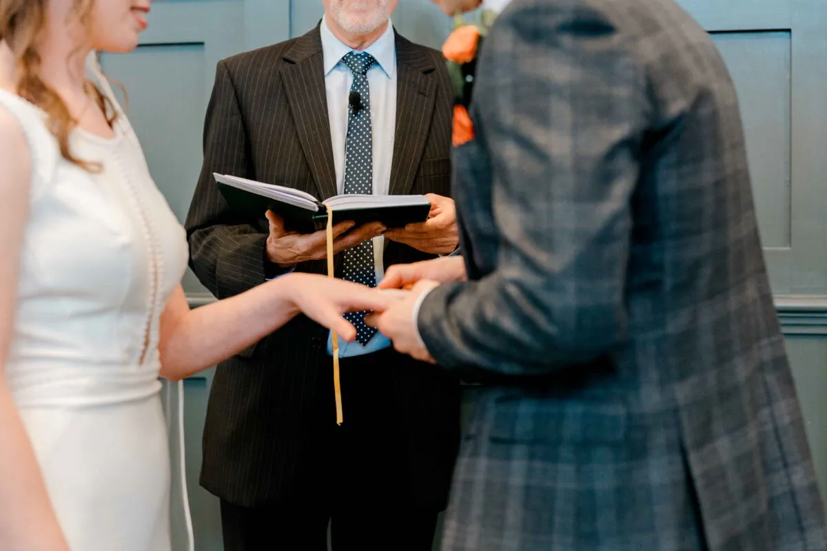The groom gently places a wedding ring onto the bride's finger while an officiant holds a ceremony book in a panelled room.