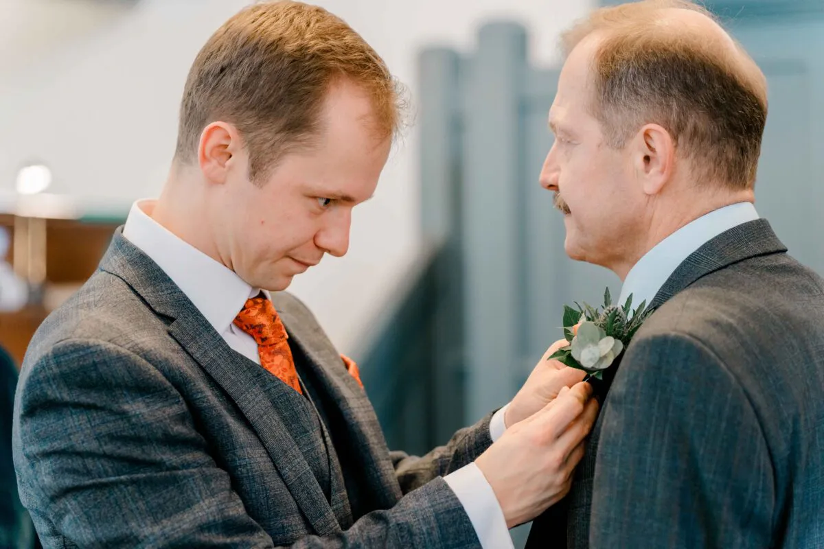 A groom in a grey suit and orange patterned tie carefully pins a floral buttonhole onto another man's lapel during wedding preparations.