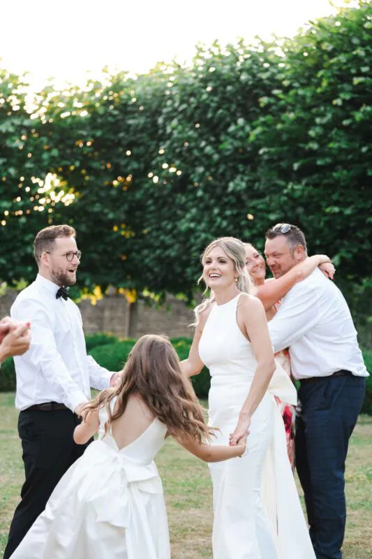 A joyous bride and groom laugh and hold hands outdoors in a sunlit garden alongside a dancing flower girl and cheering wedding guests.