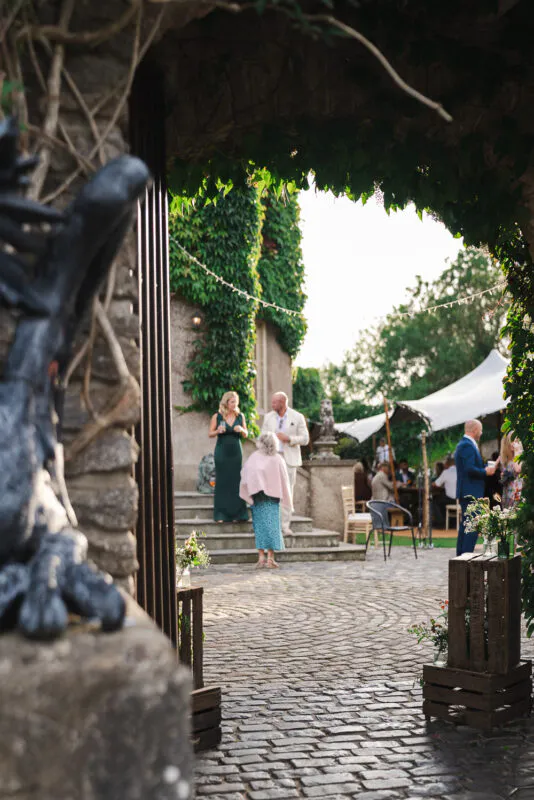 Wedding guests mingle in a sunlit cobbled courtyard, viewed through an ivy-draped stone archway with a marquee set up in the background.